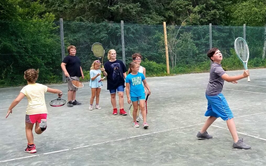 Color photo of EdGE students practicing tennis during a learning clinic. They wear shorts and t-shirts while swinging tennis rackets.