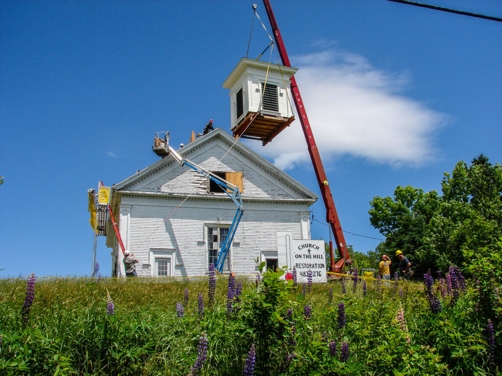 Color photo of a crane putting on a steeple on a Cherryfield Church. It is a clear day with lupines in the foreground.