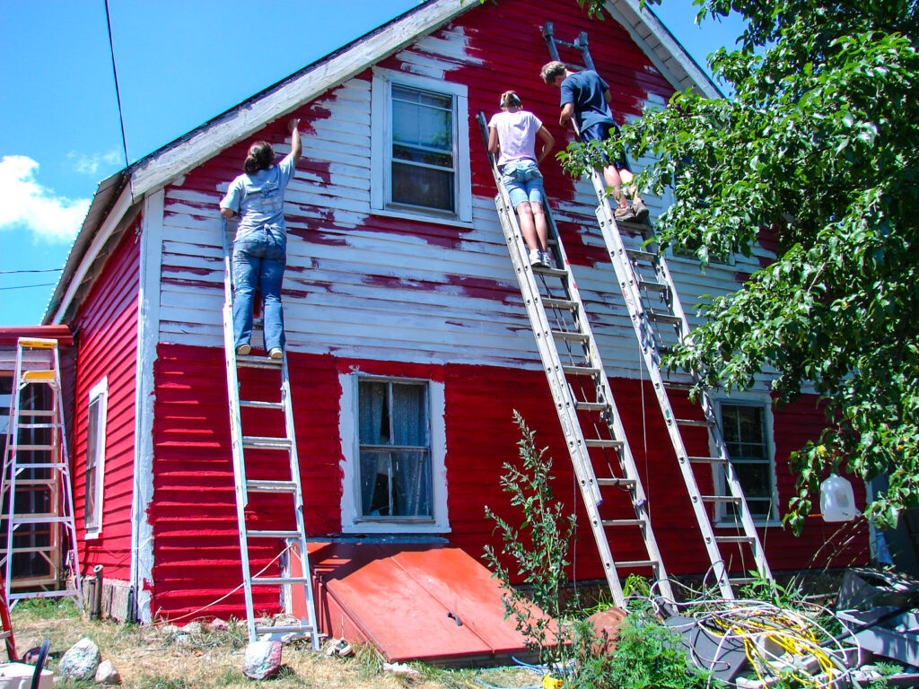 Color photo of Housing Rehab program volunteers painting a white house red on a clear day