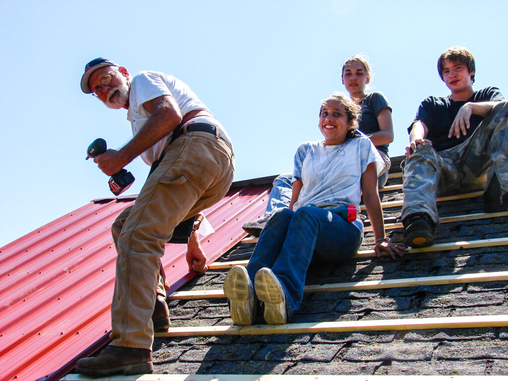 Housing Color photo of rehab program volunteers work on a red metal roof against a blue sky