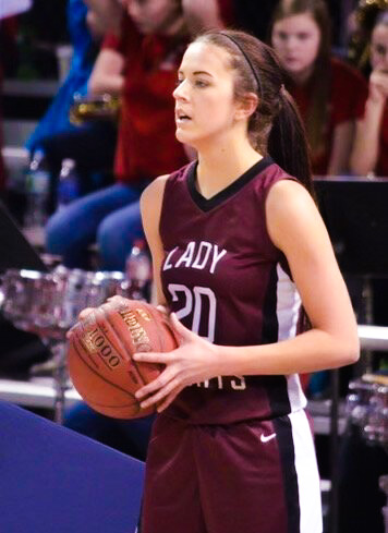 Color photo of Kelli Kennedy on a basketball court gripping a ball. She stands at an angle to the camera, her brown hair tied back and wears a jersey uniform.