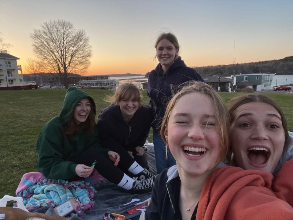 Color photo of five students laughing as they sit on the grass during a sunset.