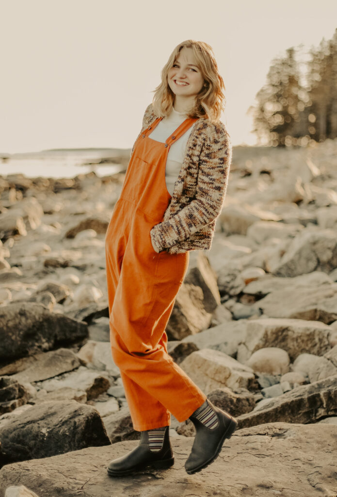 Color photo of a female student smiling and wearing overalls as she stands on the rocky shore with ocean and sky in the background.
