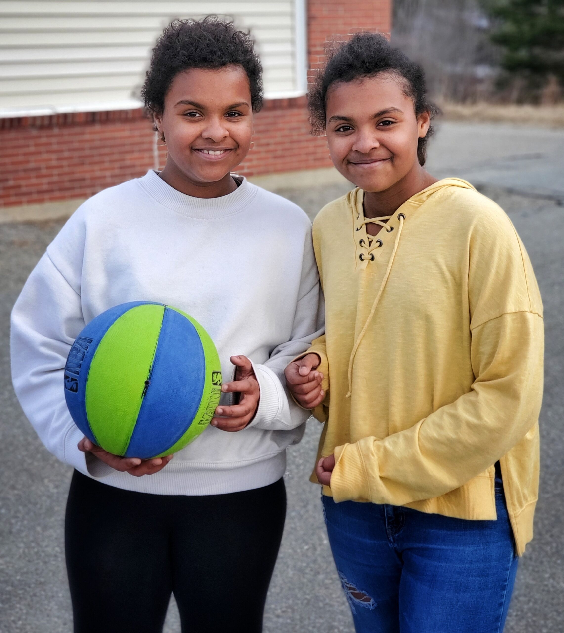 Color photograph of two pre-teen girls on a school playground. They have brown skin and are smiling. One holds a basketball.