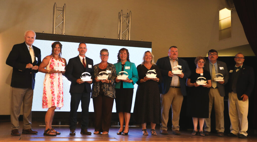 Color photo of the Downeast Education Partners, nine in all, stand on stage with their awards in hand. President John Zavodny and retired Executive Director Gary DeLong stand on either side of the group.