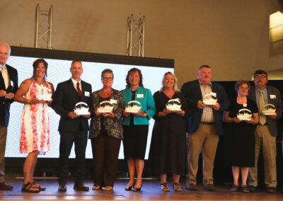 Color photo of the Downeast Education Partners, nine in all, stand on stage with their awards in hand. President John Zavodny and retired Executive Director Gary DeLong stand on either side of the group.