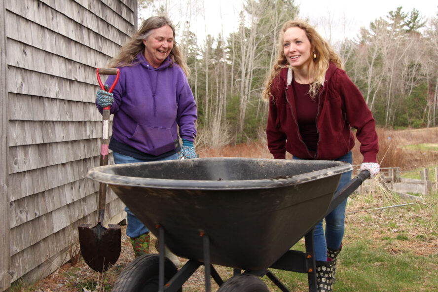 A color photo of two white women smiling pushing a wheelbarrow