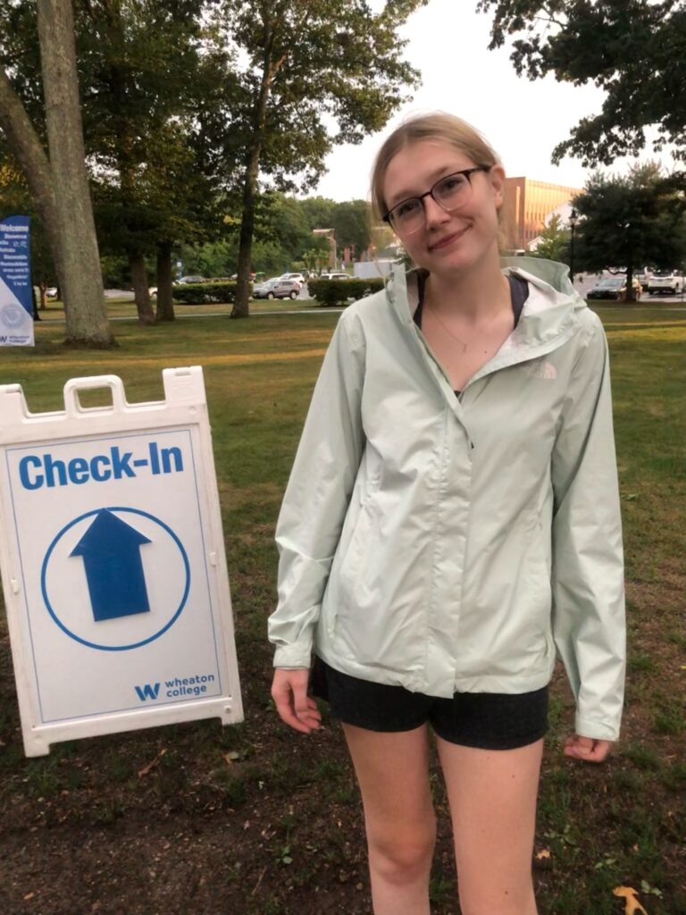 A color photo of a college student standing in front of a "check-in" sign