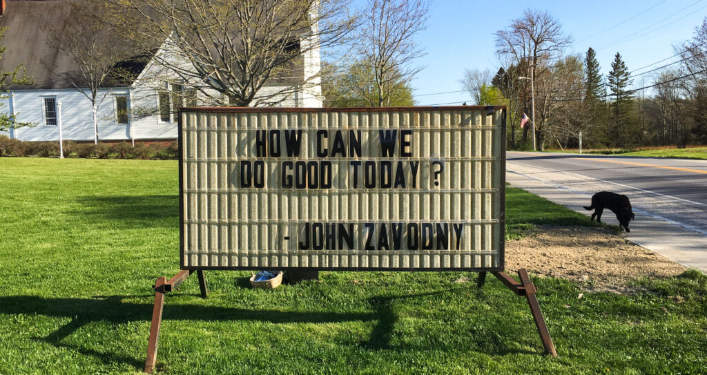 A color photo of a sign that says "How can we do good today? - John Zavodny"