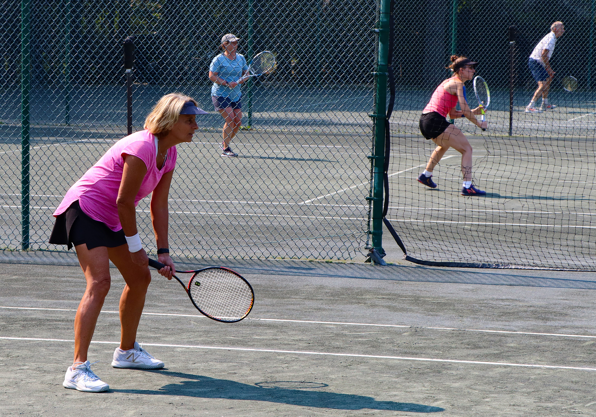 Color photo of three female tennis players on a clay court under the sun. They are in an athletic stance and have tennis rackets in hand.