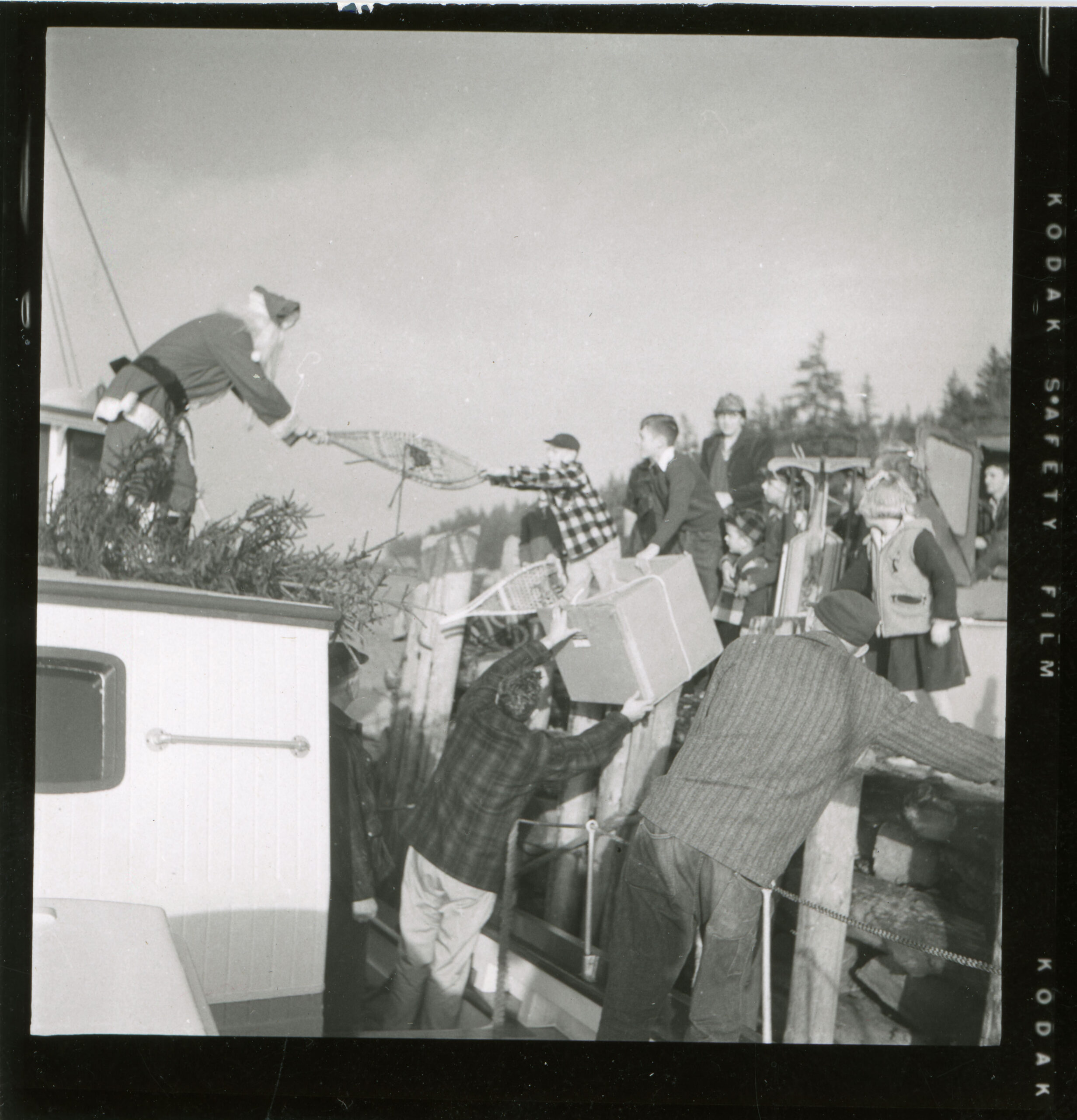 A black and white photo of a man dressed as Santa handing gifts off of a boat