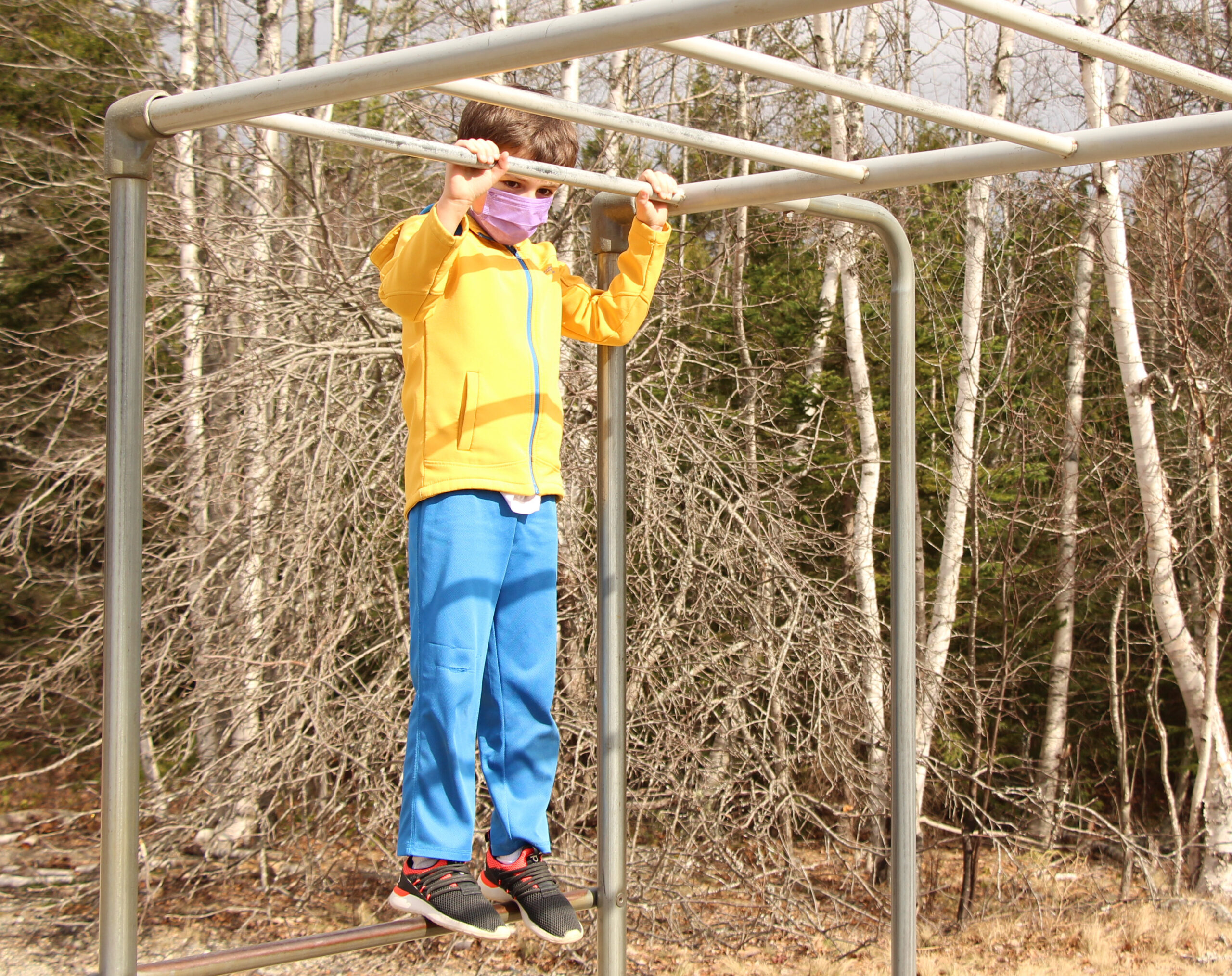 Color photo of a white child wearing a bright yellow jacket stands on monkey bars at an elementary school. Trees are in the background and it is spring.