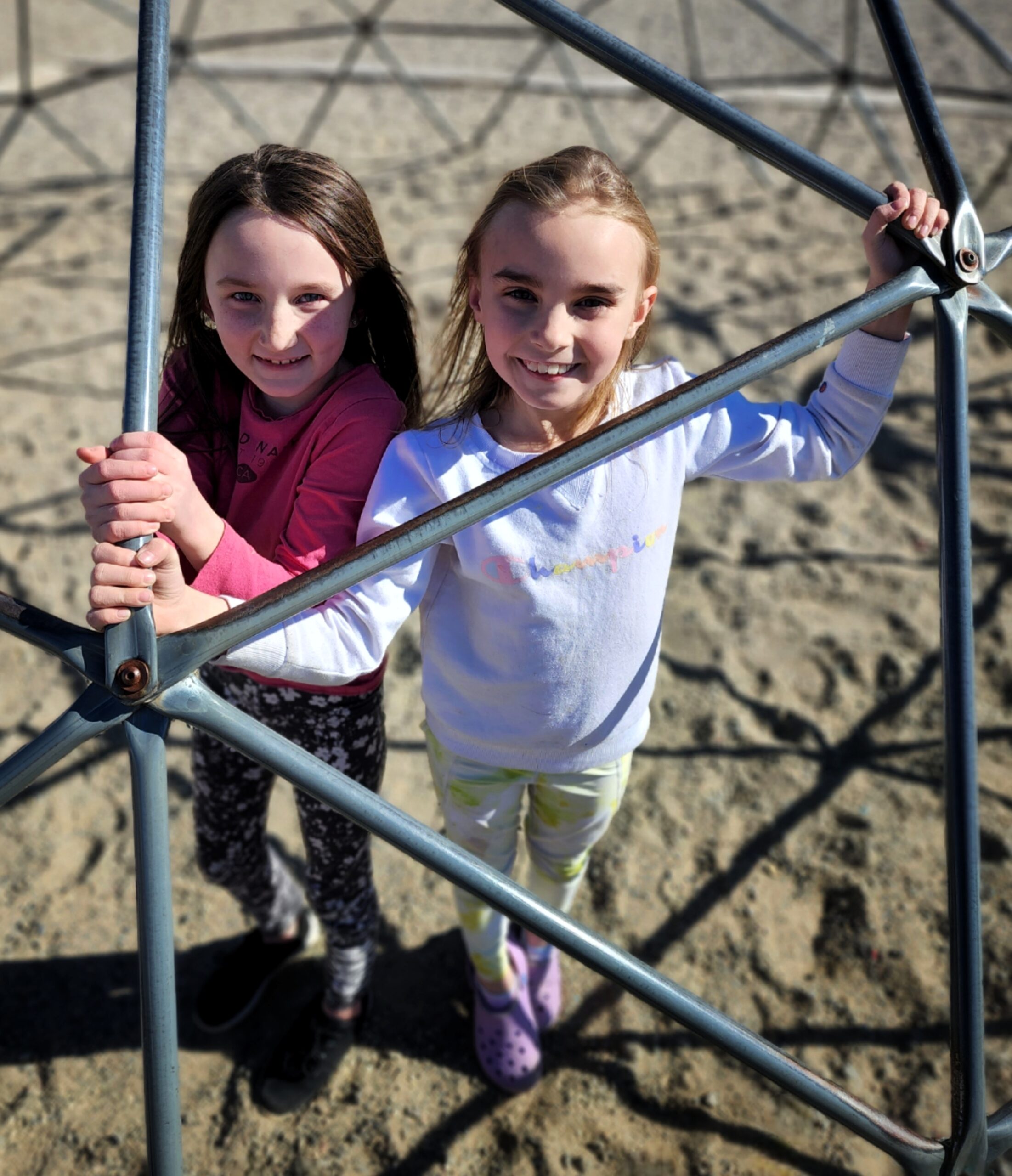 Color photo of two white female children stand inside a play area dome. It is a sunny day and they smile at the camera.