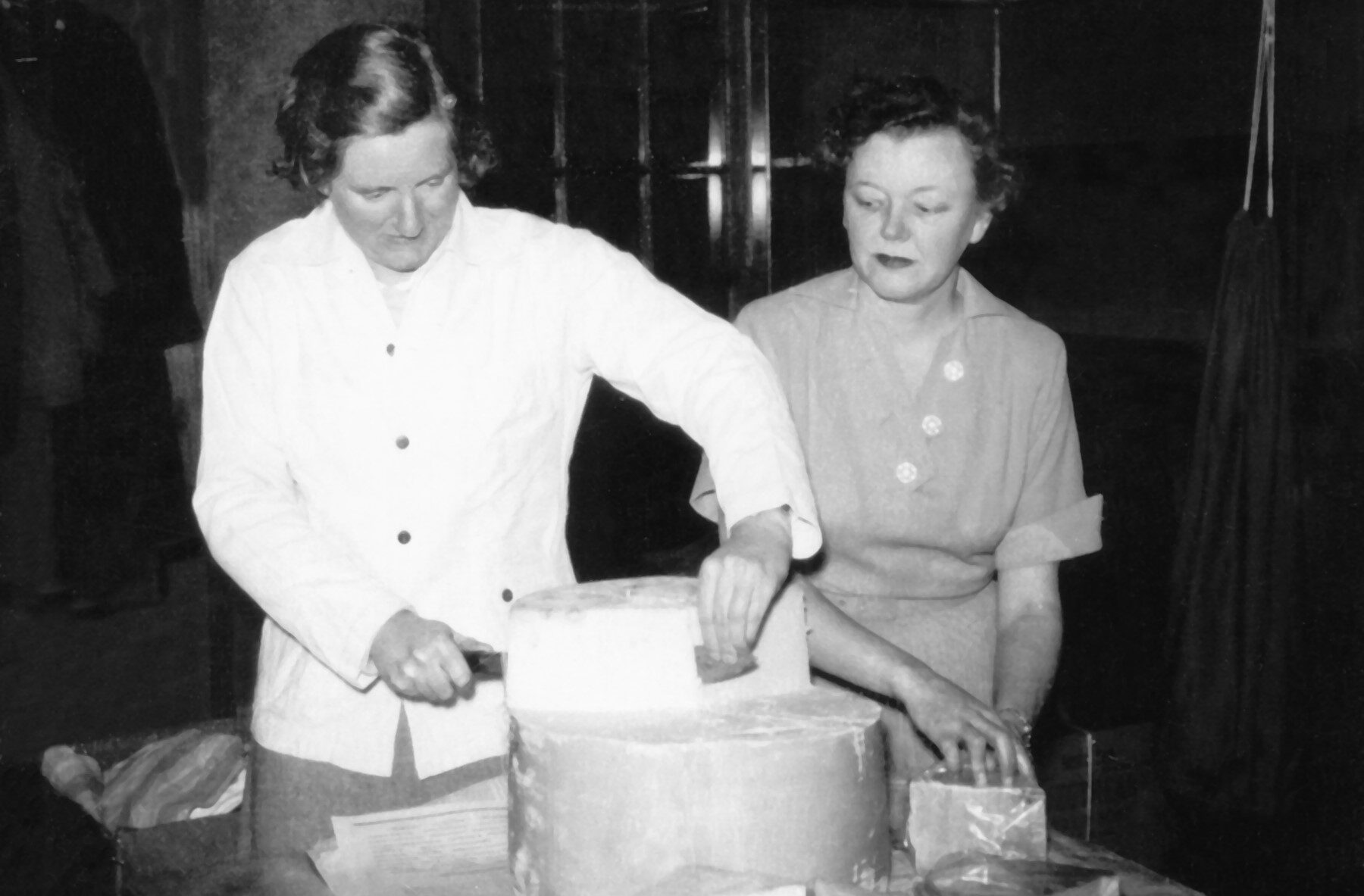 A black and white photo of two women cutting cheese