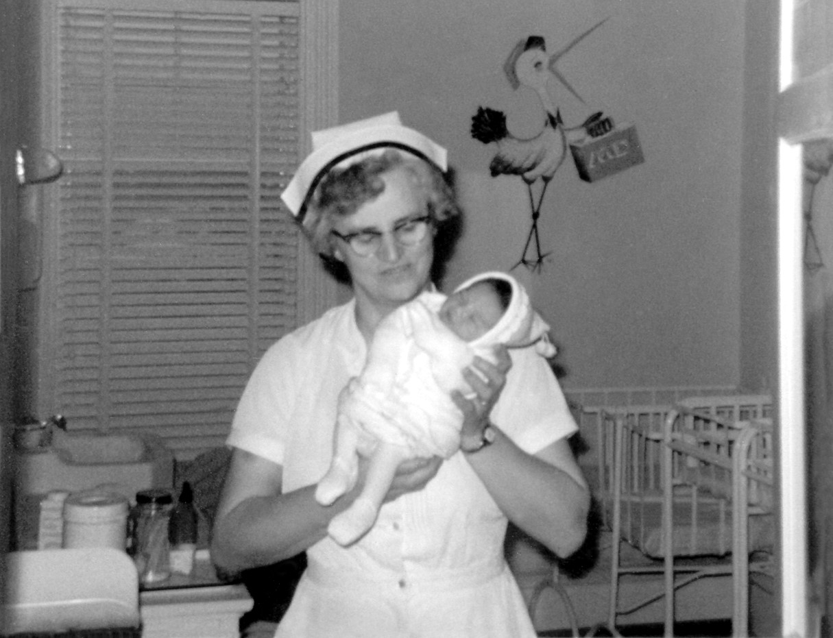 A black and white photo of a nurse holding a baby in front of a painting of a stork