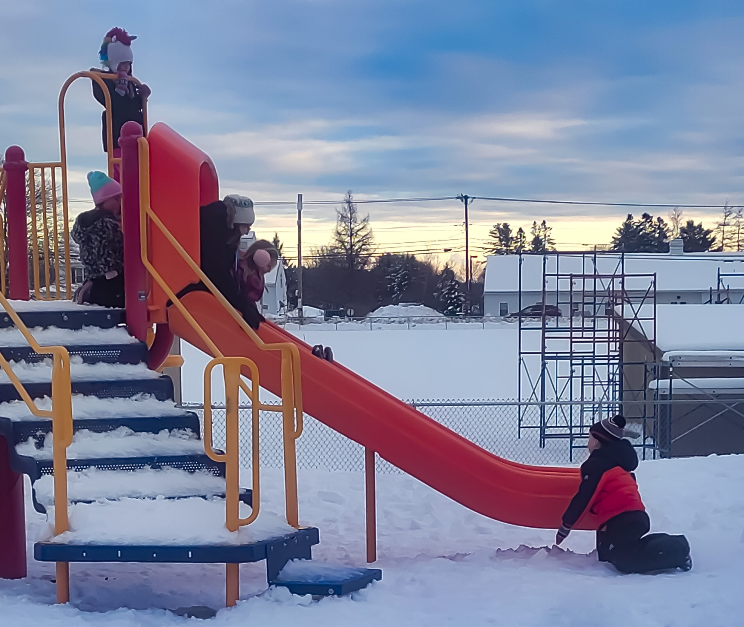 Color photo of four children playing on a snow-filled slide play structure during winter.