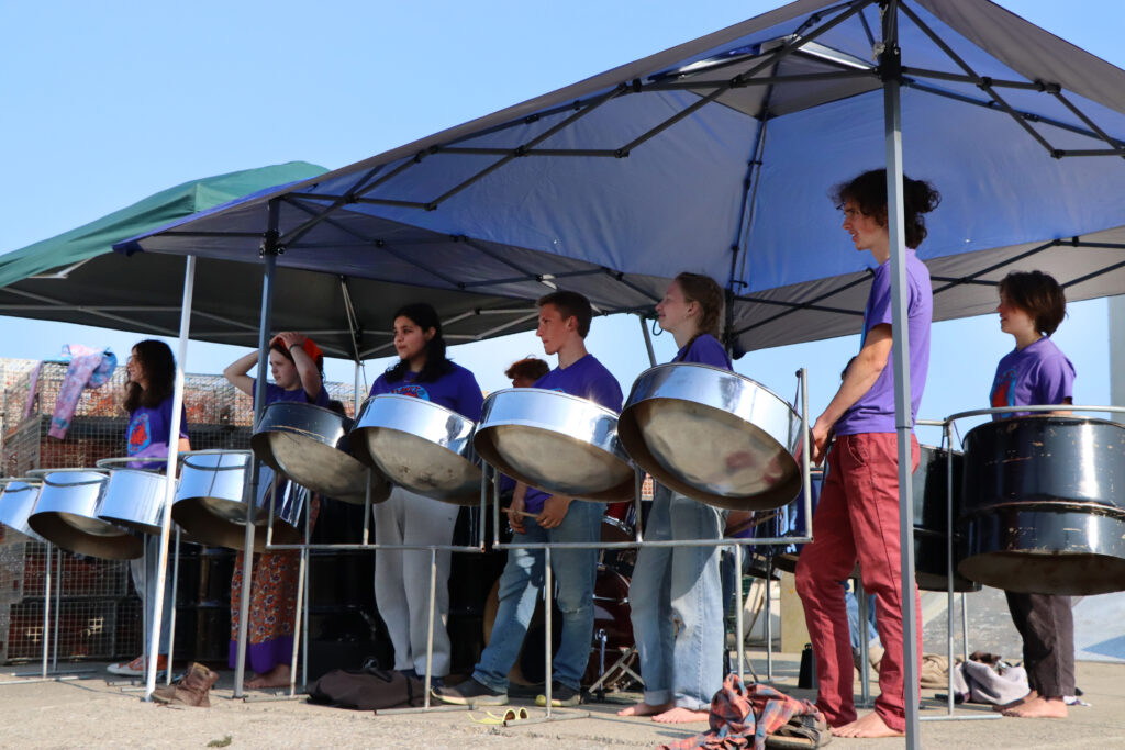 Color photo of teenagers stand behind steel pans. They wear bright purple t-shirts and are beating the pans with mallets.