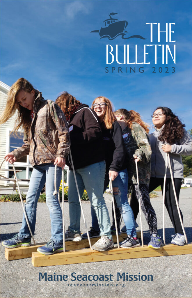 Color photo of five female teenagers standing on beams of wood and stepping together for a team-building exercise. They are smiling and concentrating.