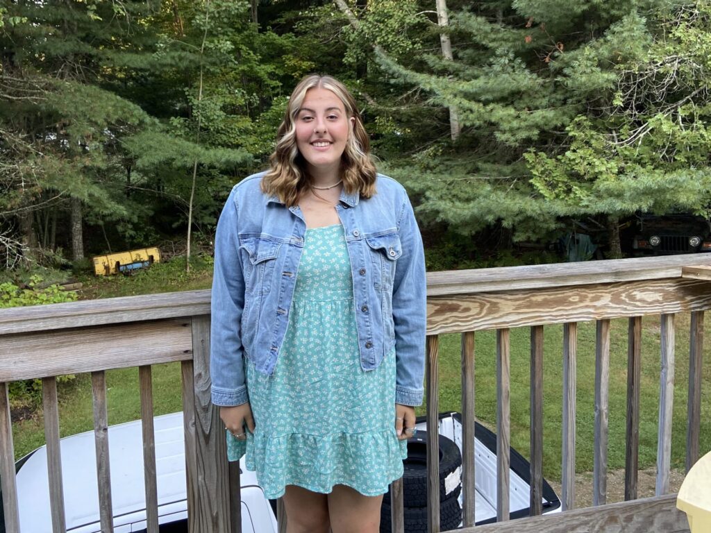 A young woman smiles at the camera while wearing a jean jacket and green dress