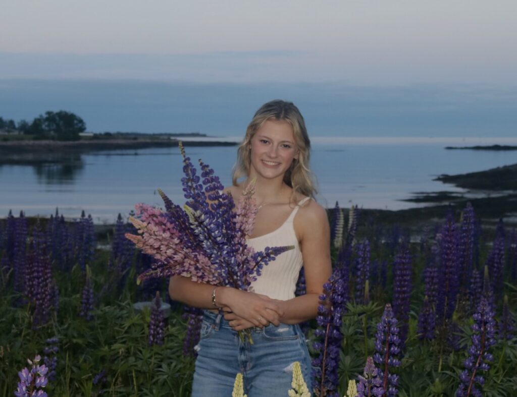 A smiling young white woman is posed in a field of lupin, holding more in her arms.