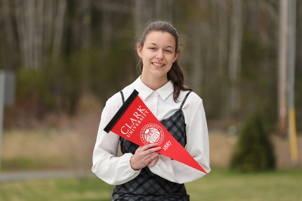 Photo of a young white woman standing in a library holding a pennant that says Clark University