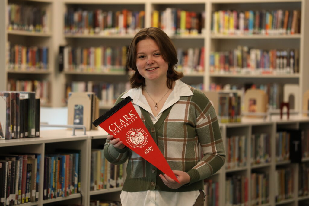 Photo of a young white woman standing in a library holding a pennant that says Clark University