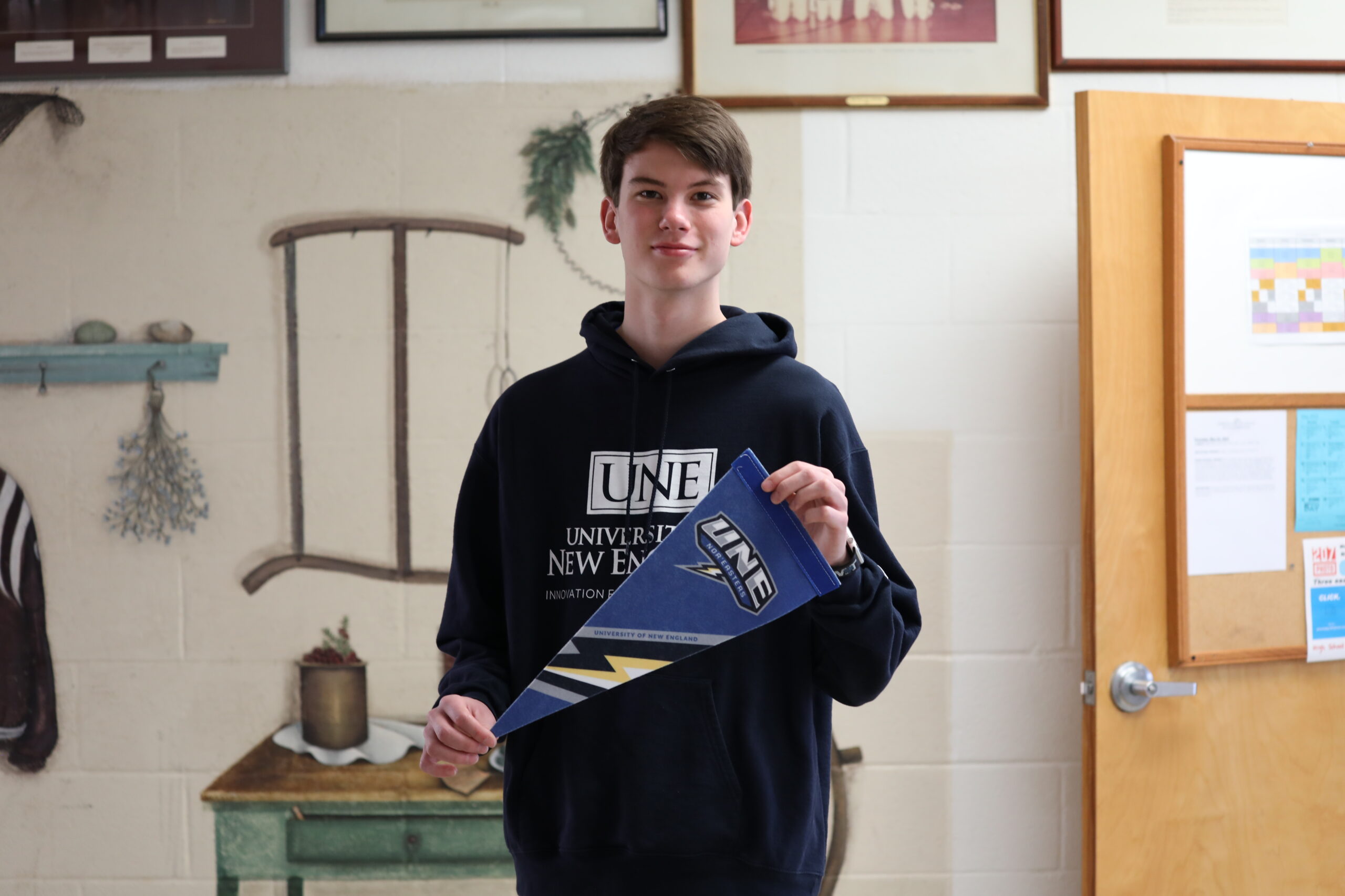 A photo of a young smiling white man holding a college pennant that says UNE