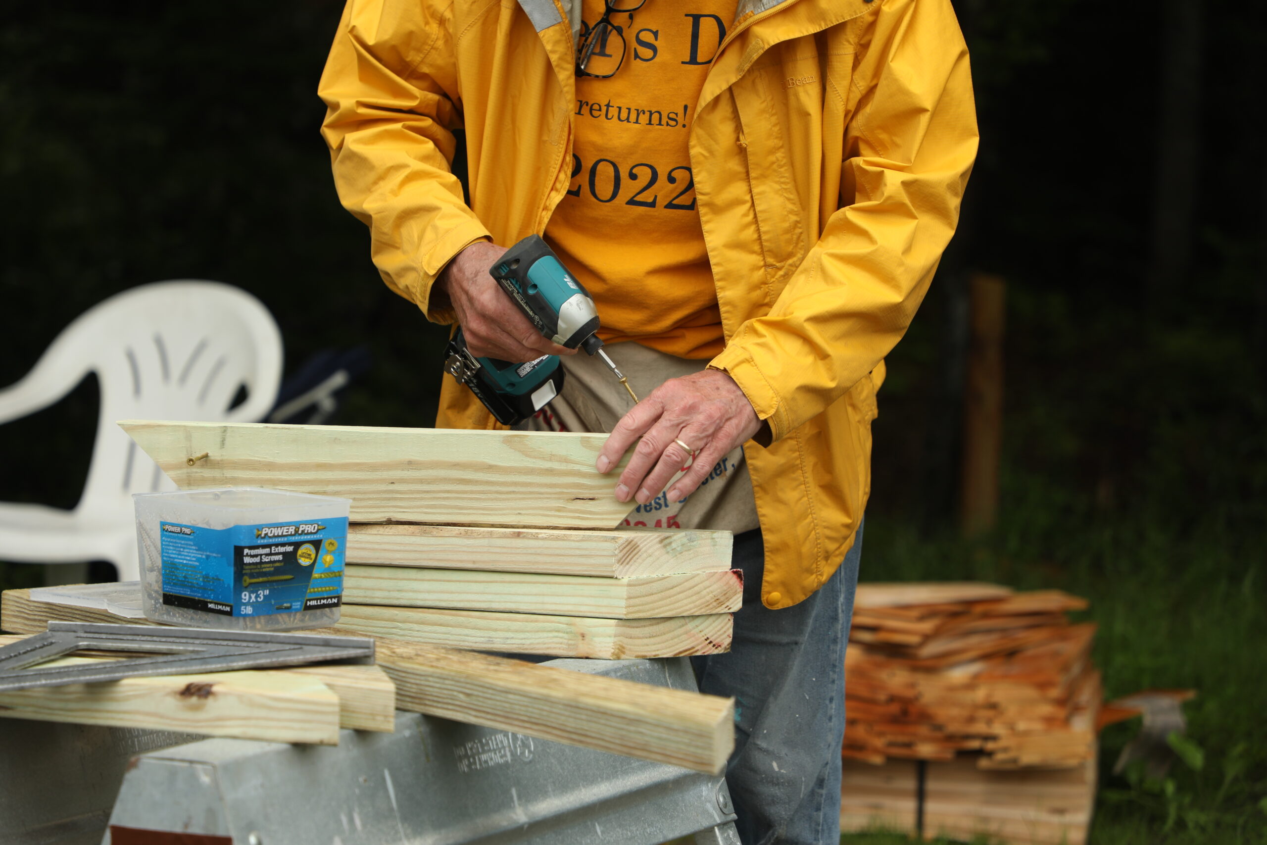 A close up of a man drilling into a piece of wood. He is wearing a yellow rain jacket and stands in front of a saw horse.