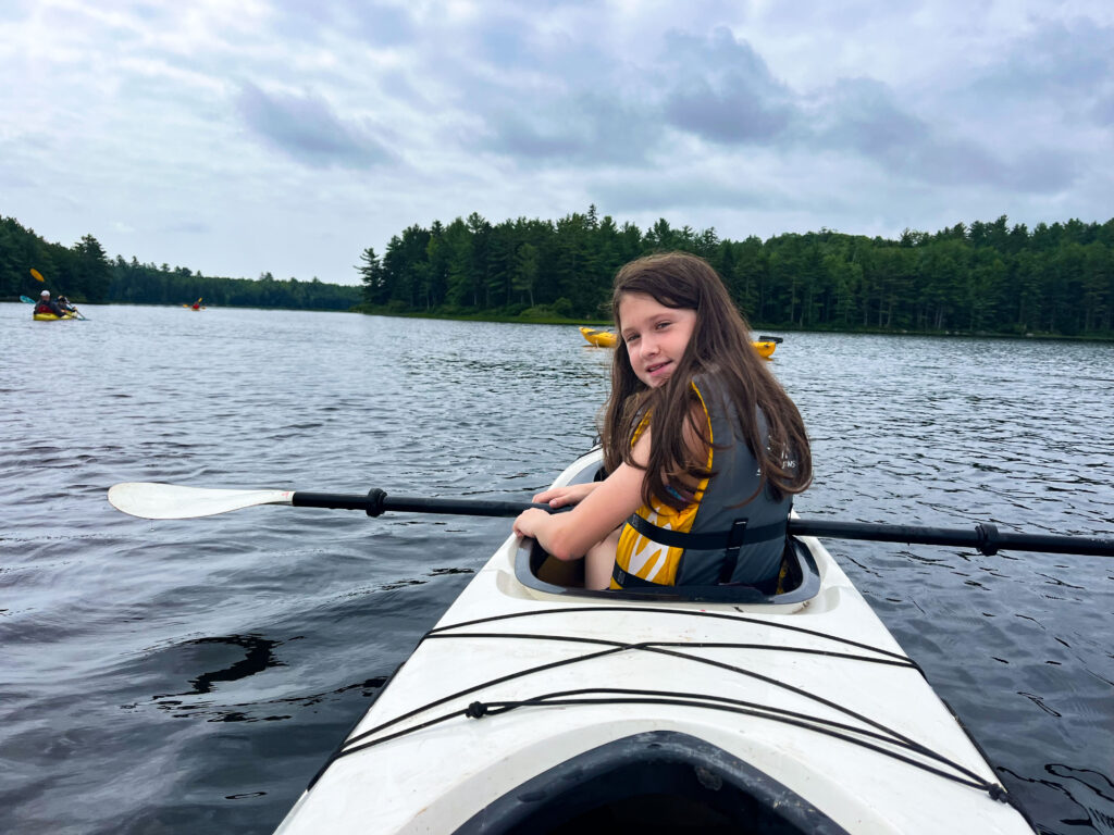 A color photo of a young white girl looking back at the camera and smiling while paddling a white kayak