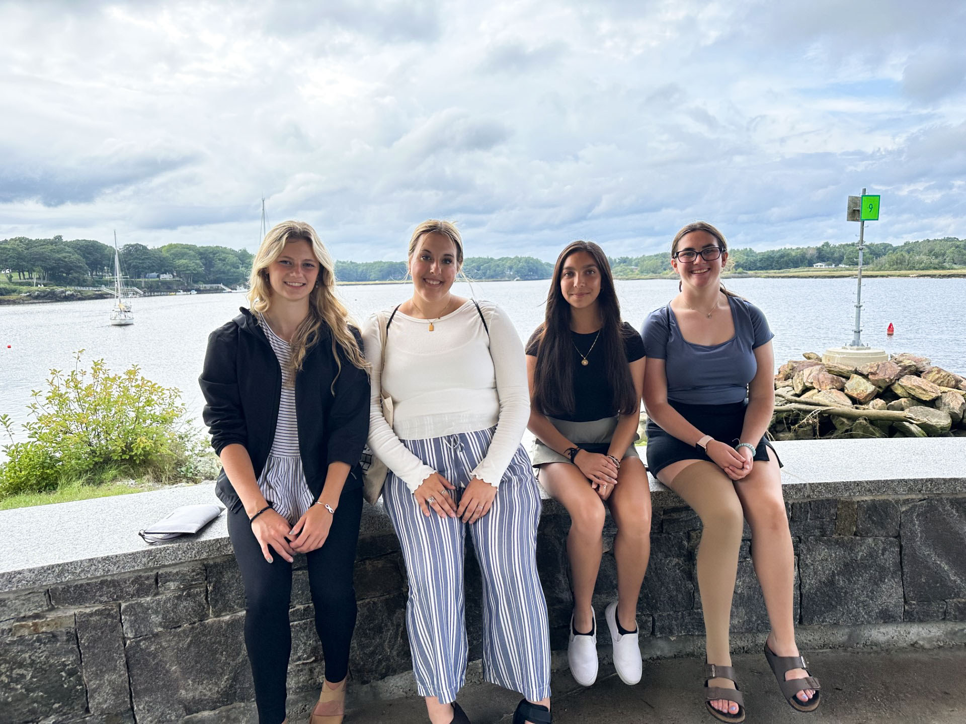 A photo of four young women posing in front of the ocean. They are sitting on a rock wall