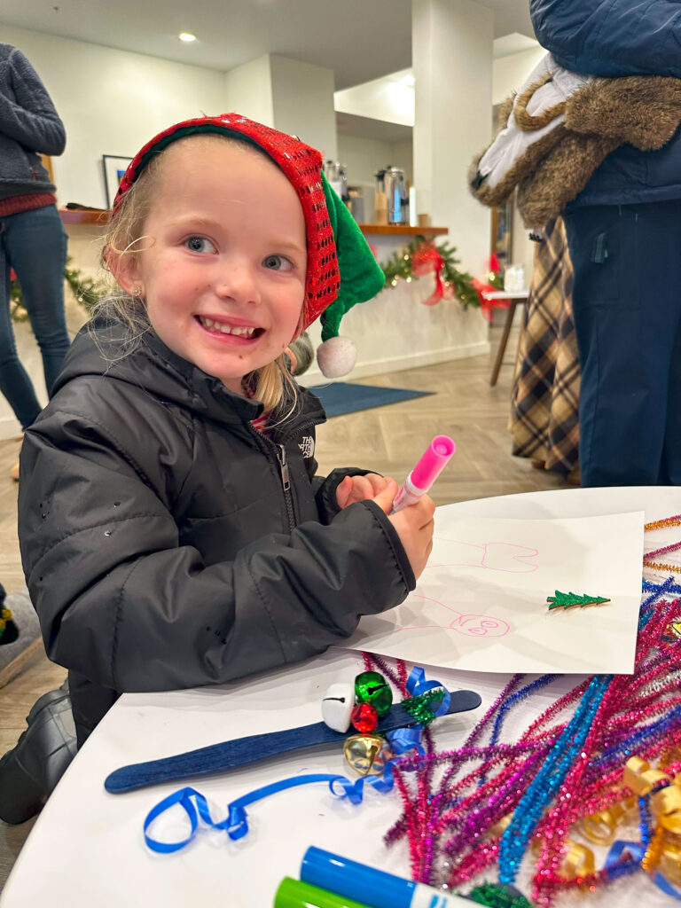 A young girl wearing a Christmas hat smiles at the camera while coloring