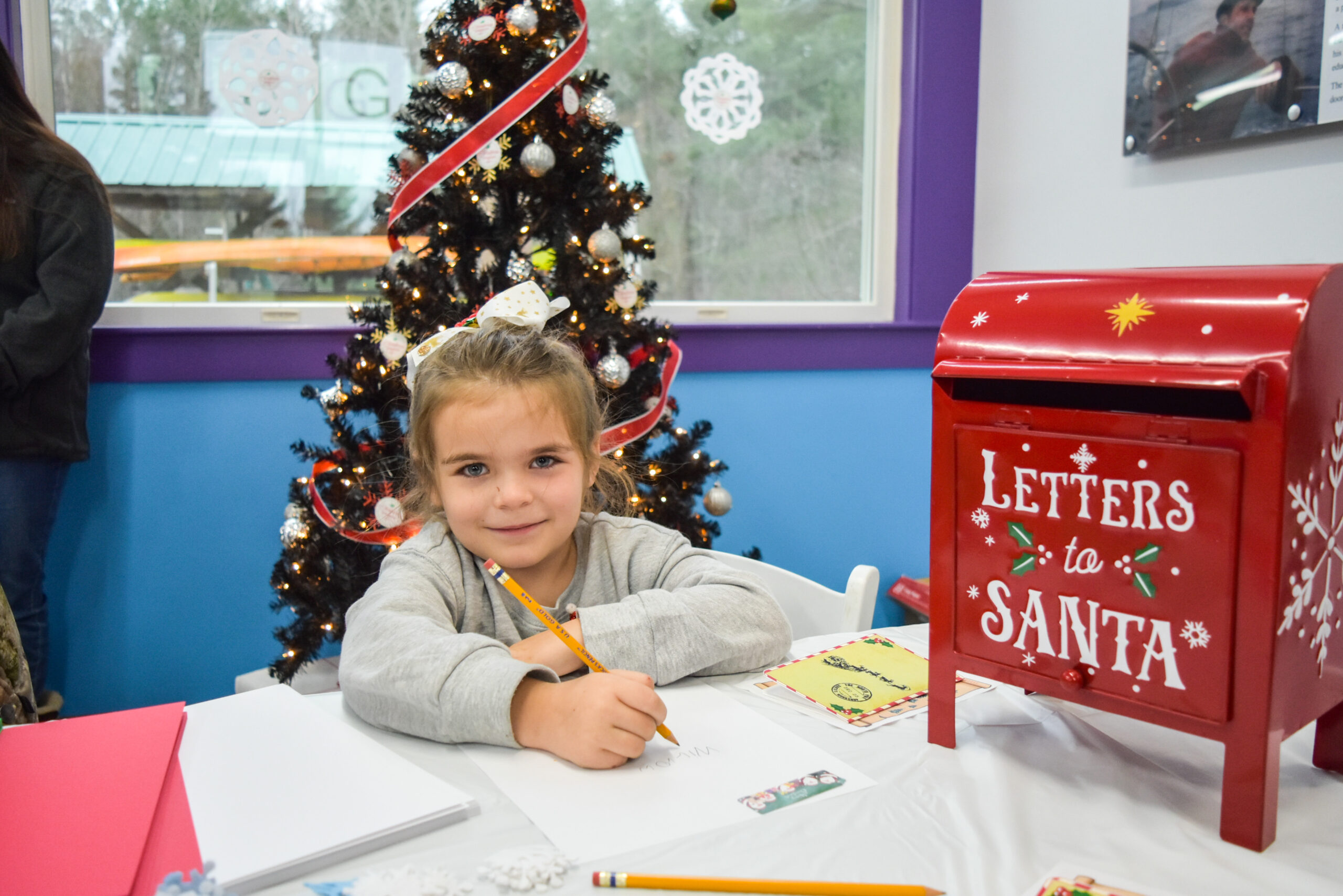 A young girl looks at the camera in front of a Christmas tree. She is writing on a piece of paper and is next to a mailbox that reads "Letters to Santa"