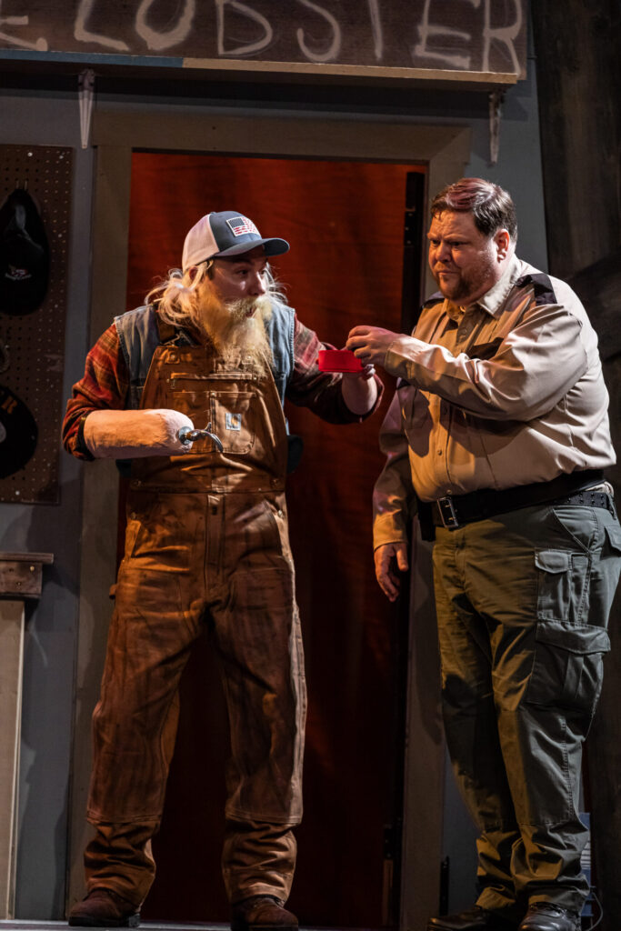 Two white men dressed as a lobsterman and a police officer examine a red thermos cup.