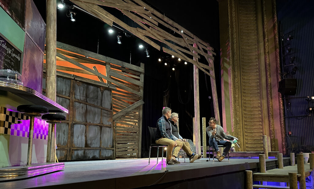 Three white men sit on a theatre stage and converse.