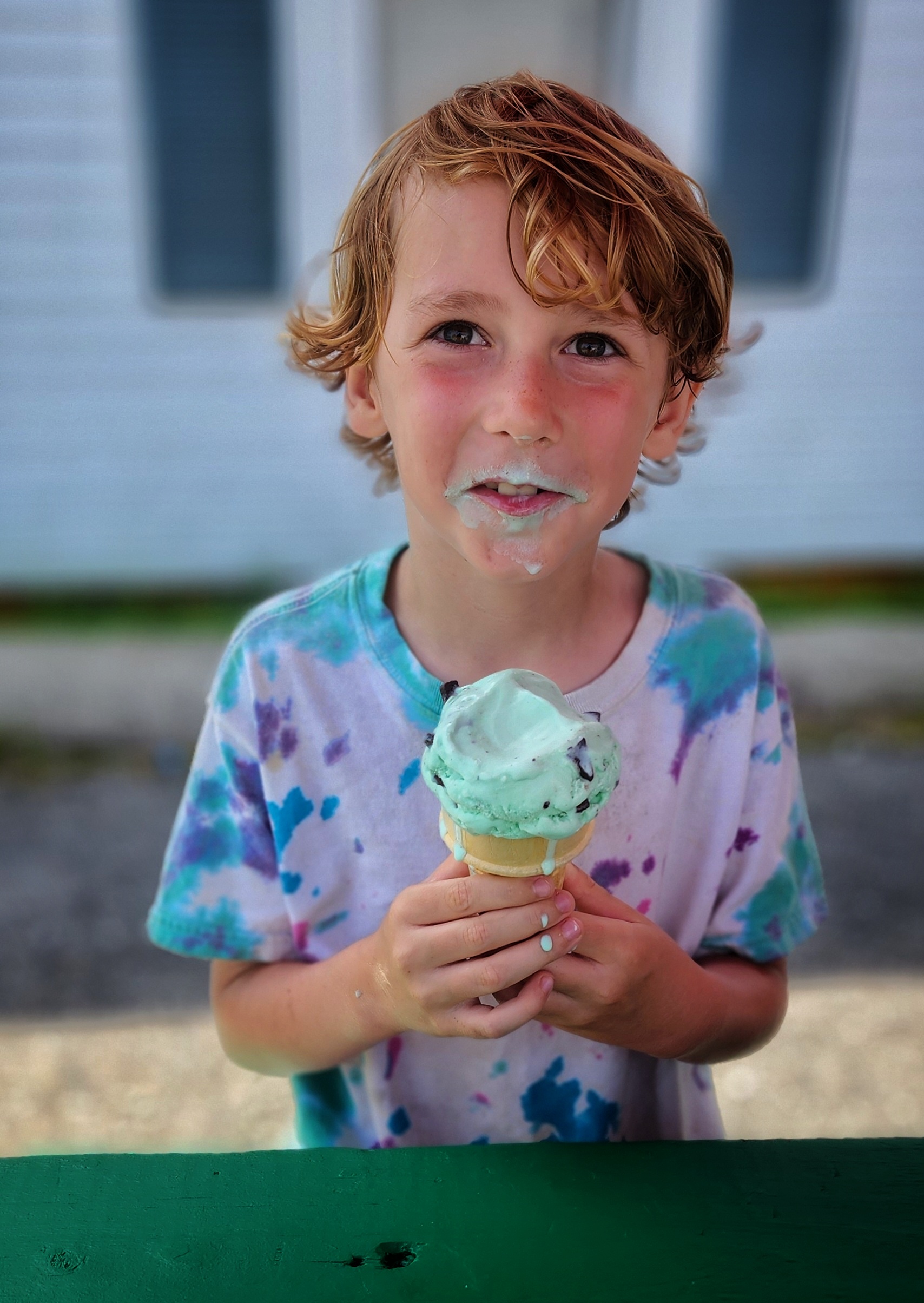 A color photo of a young white boy eating ice cream. He is looking at the camera and smiling with ice cream on his face. 