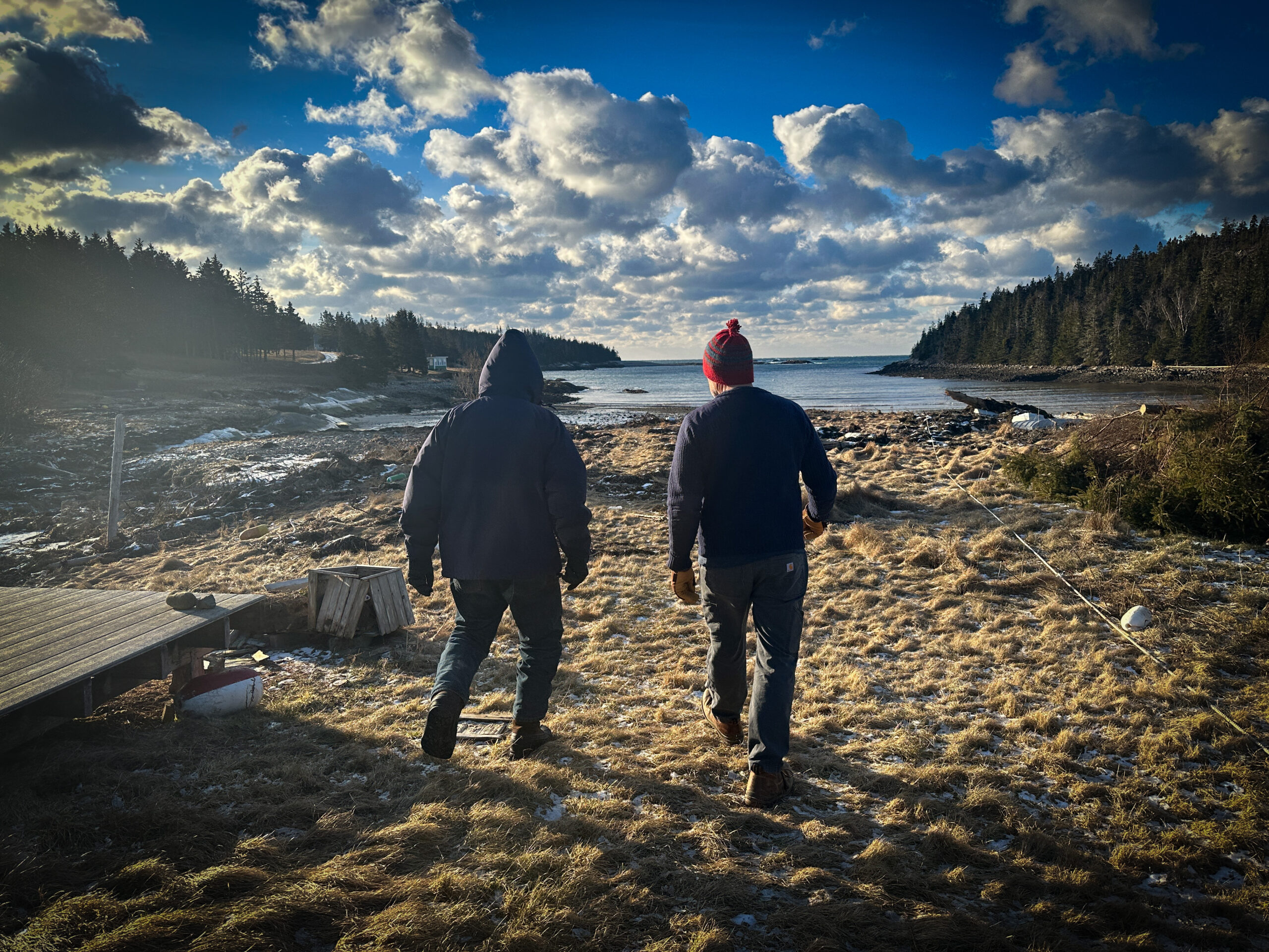 Two male figures walk across winter grass toward a beach. The sky is bright with sun and speckled with clouds. There is debris on the beach and grass caused by severe storms.