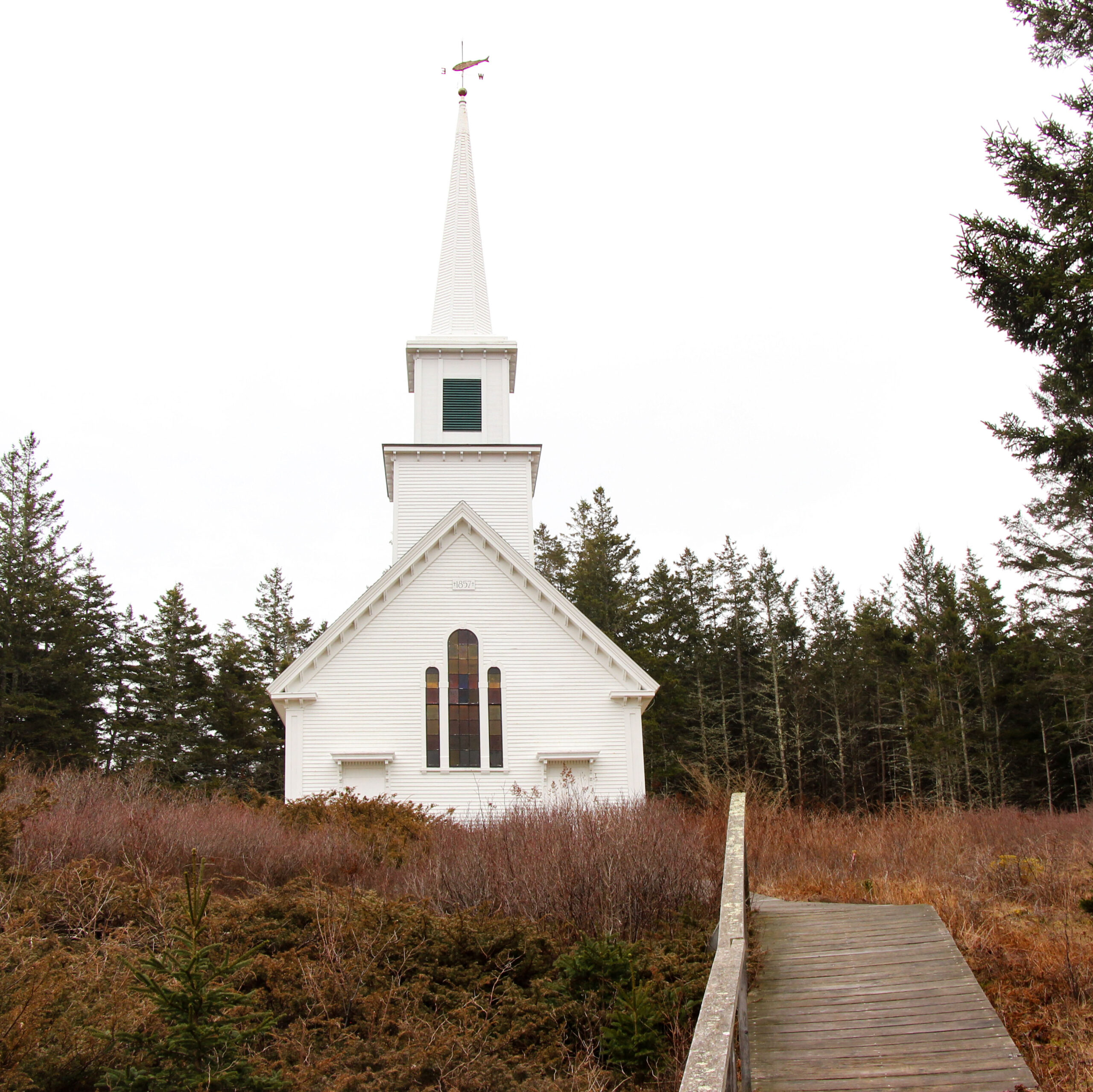 A photo of a church on a hill. It is a simple white church with a steeple and stained glass window 