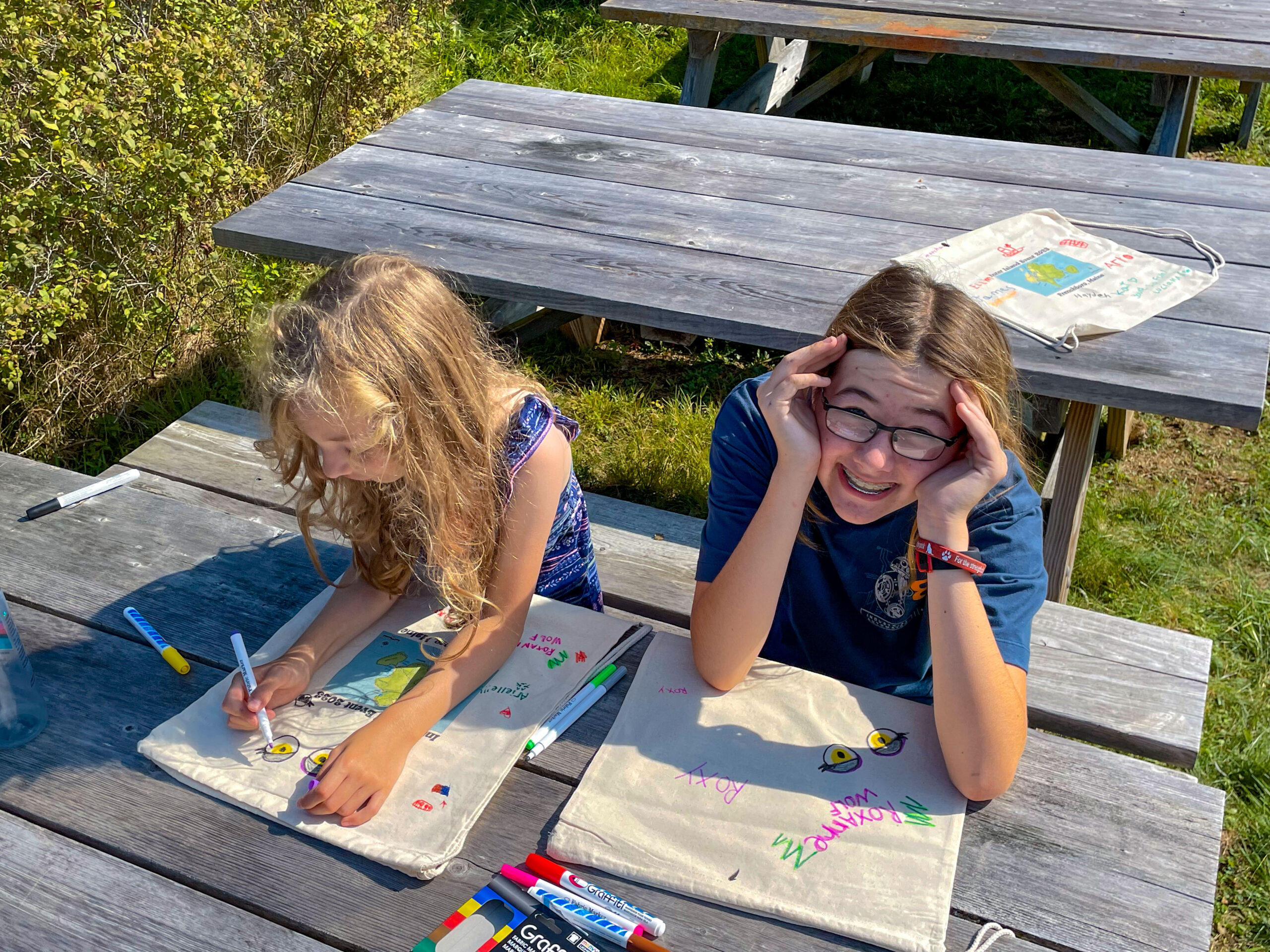 Two young female students sit at a picnic table. One looks up at the camera smiling she is shielding her eyes from the sun. 