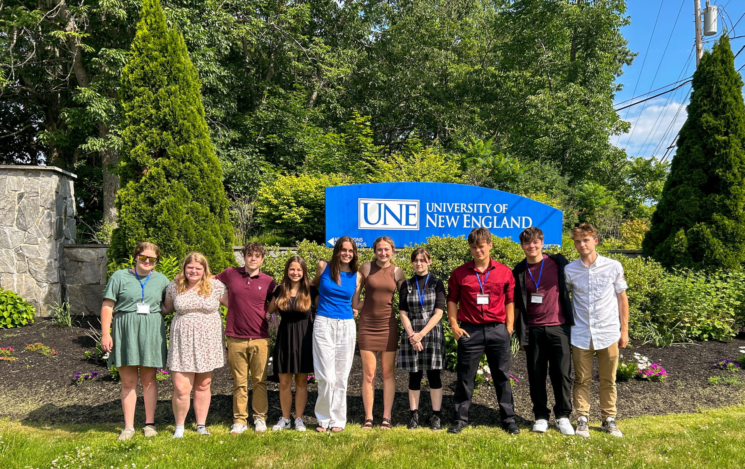 A group of smiling teenagers stand in front of a sign that reads "UNE University of New England"