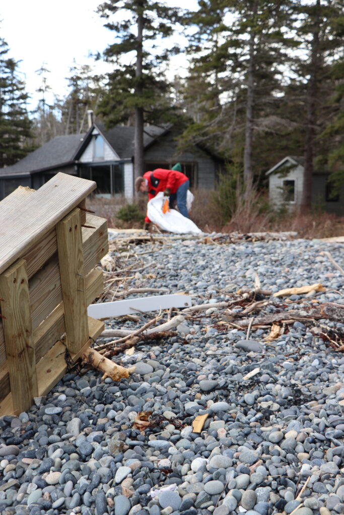 A photo of a person picking up trash on a beach. In the foreground are steps that had been washed ashore. 