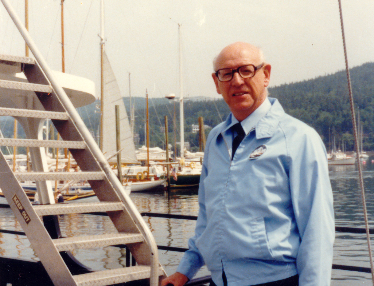 An older white man stands on a boat with the ocean behind him. He wears thick black glasses and a blue jacket.