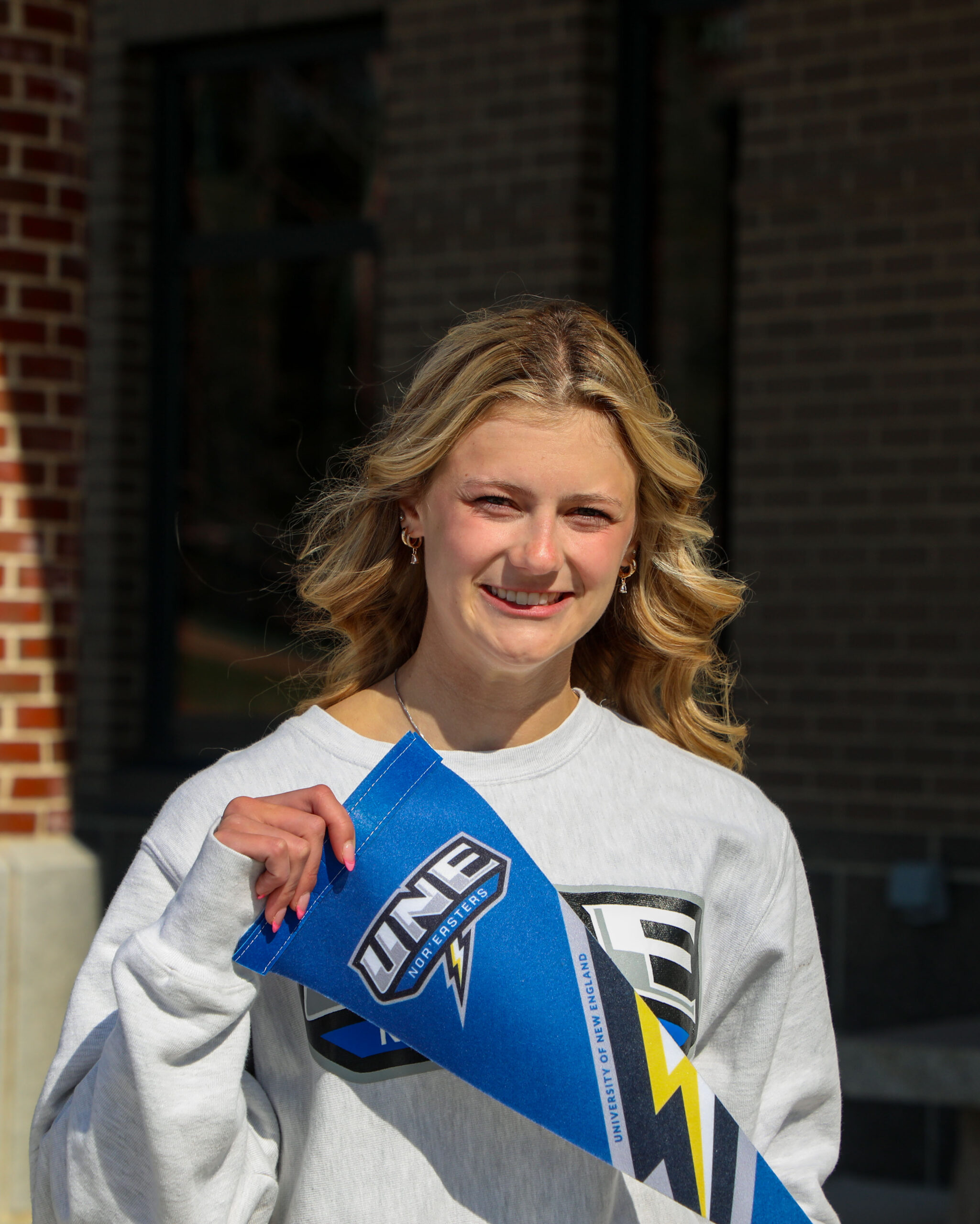 A color photo of a young woman smiling at the camera on a very sunny day. In her hands in a pennant for the University of New England.