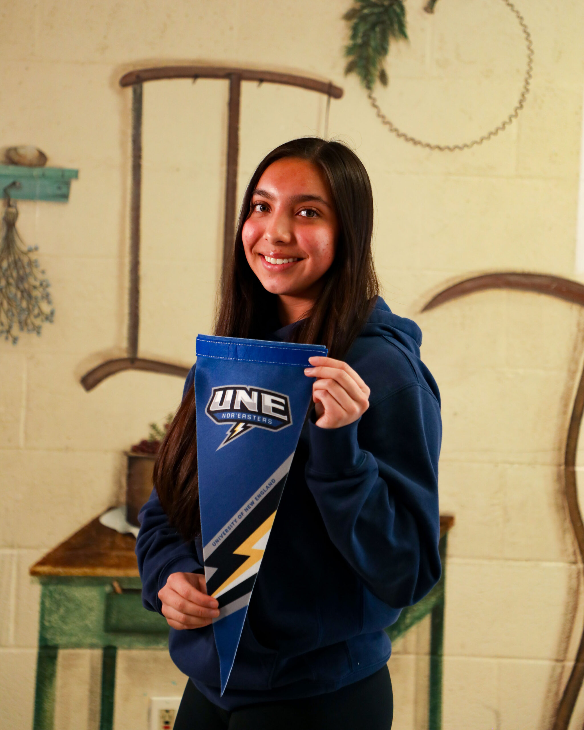 A color photo of a young woman smiling at the camera in front of a mural. In her hands in a pennant for the University of New England.