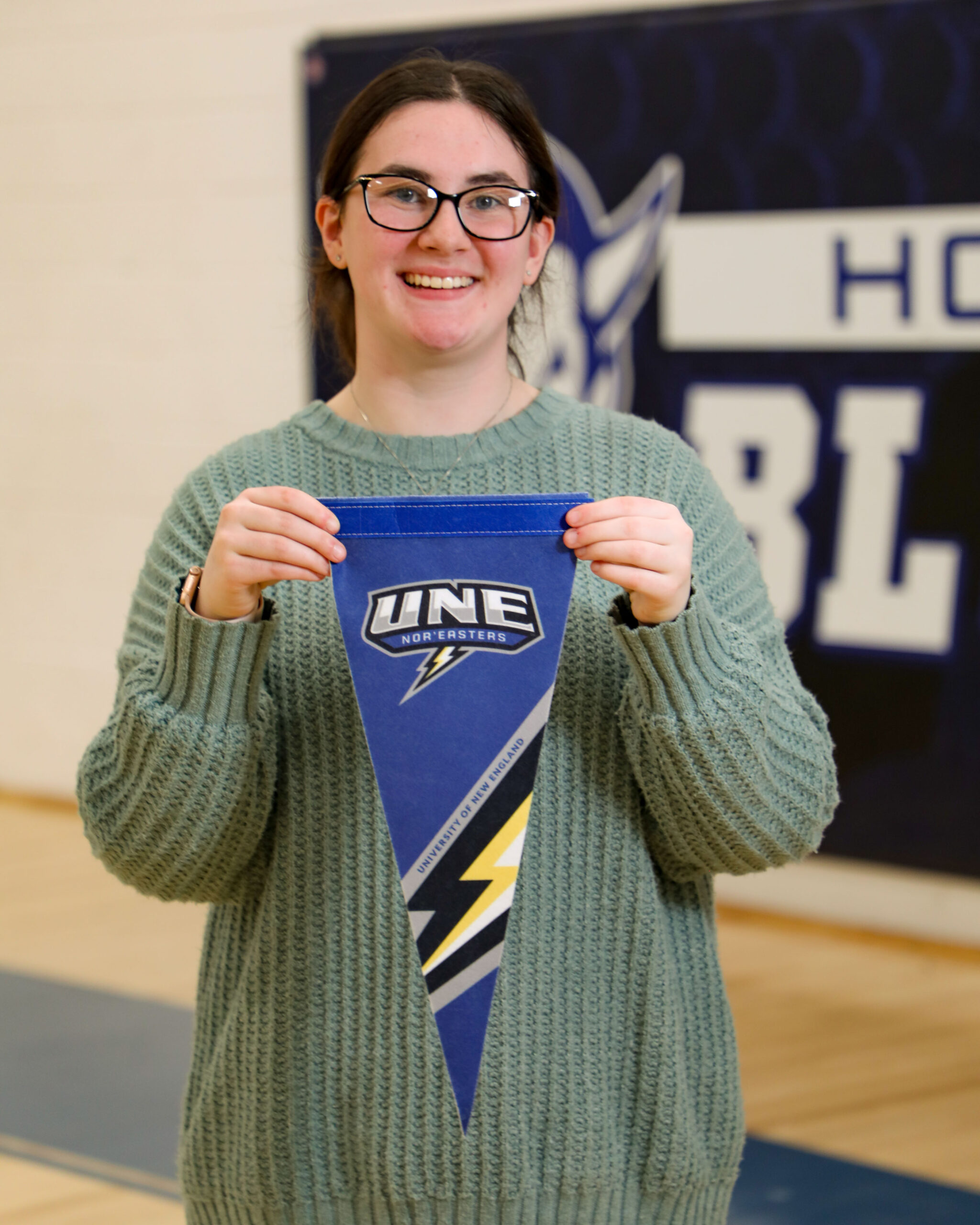 A color photo of a young woman with glasses smiling at the camera. In her hands in a pennant for the University of New England.