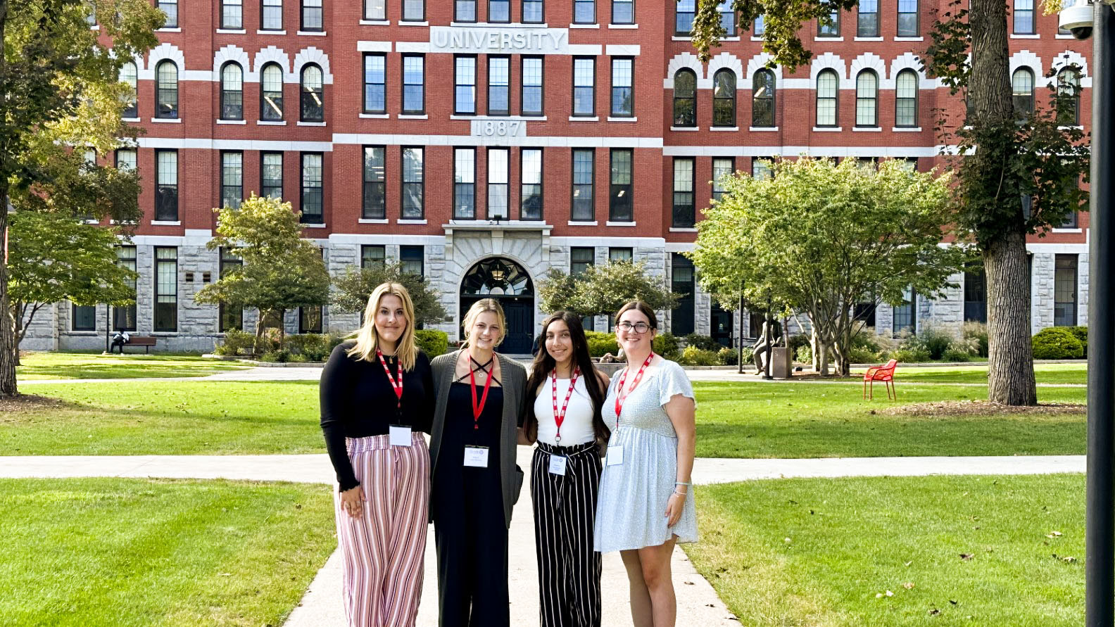 Four young women stand in front of a building that reads "Clark University" They are smiling at the camera and are all wearing bright red lanyards.