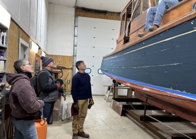 A man sits aboard a lifted boat in a repair shop and converses with five people standing on the floor below him