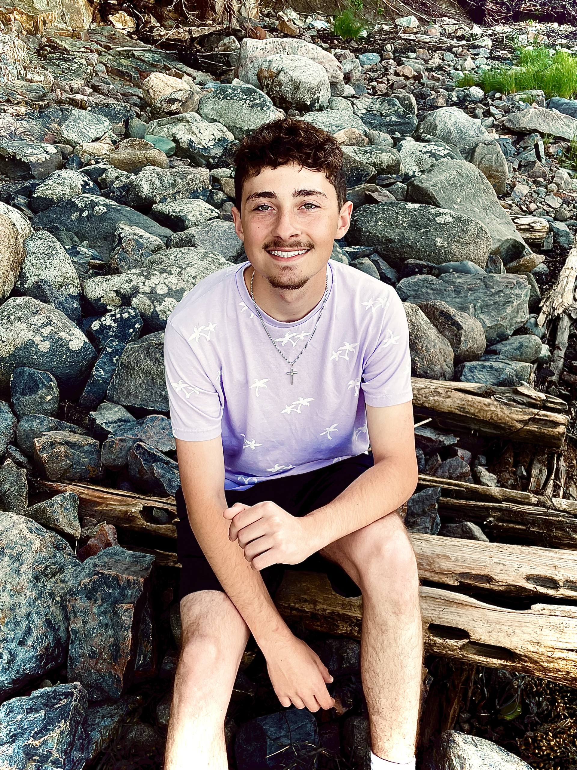 A young man smiles at the camera, he is siting on rocks at the beach.