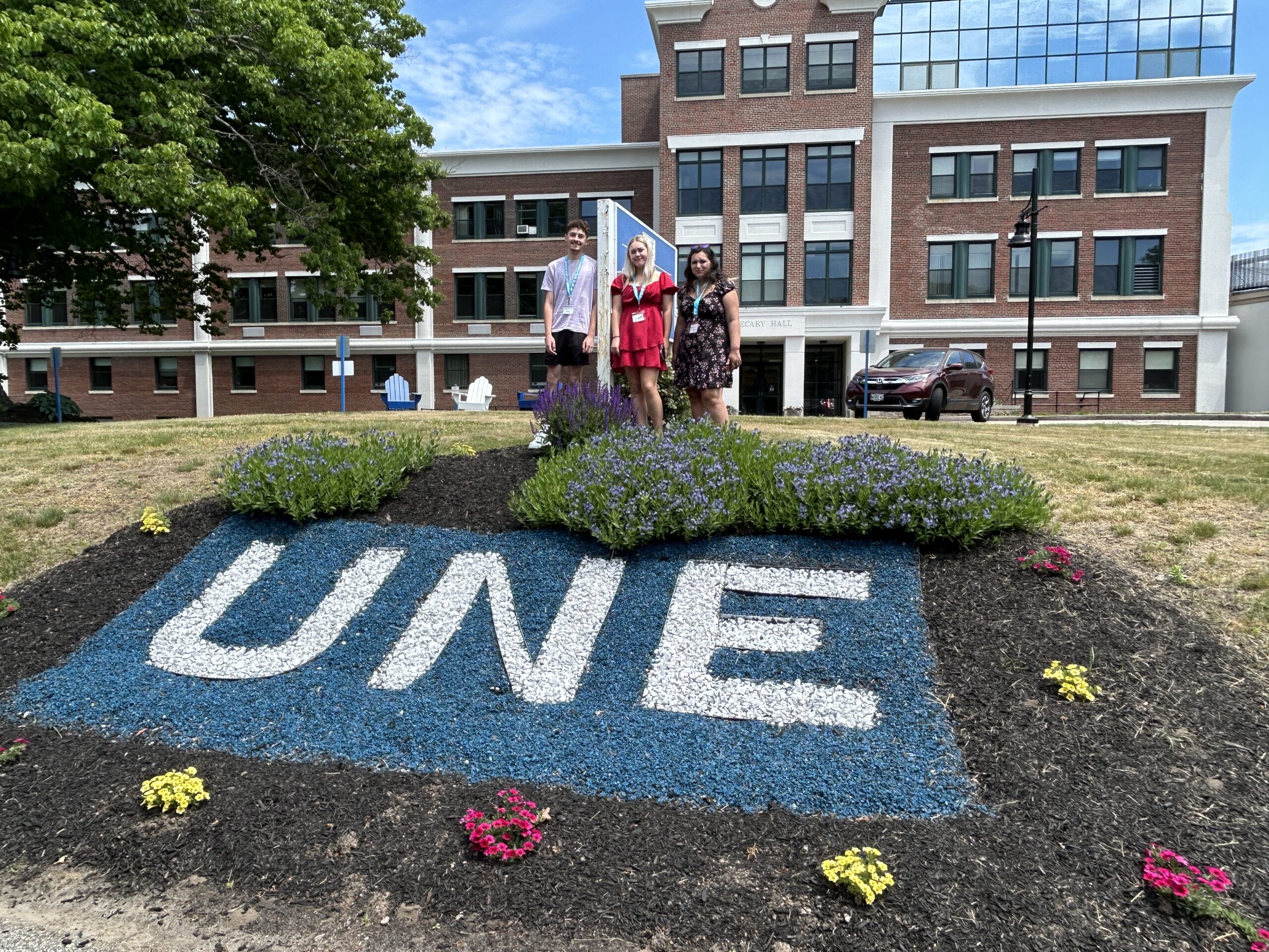 A group of smiling teenagers stand in front of a sign that reads "UNE University of New England"