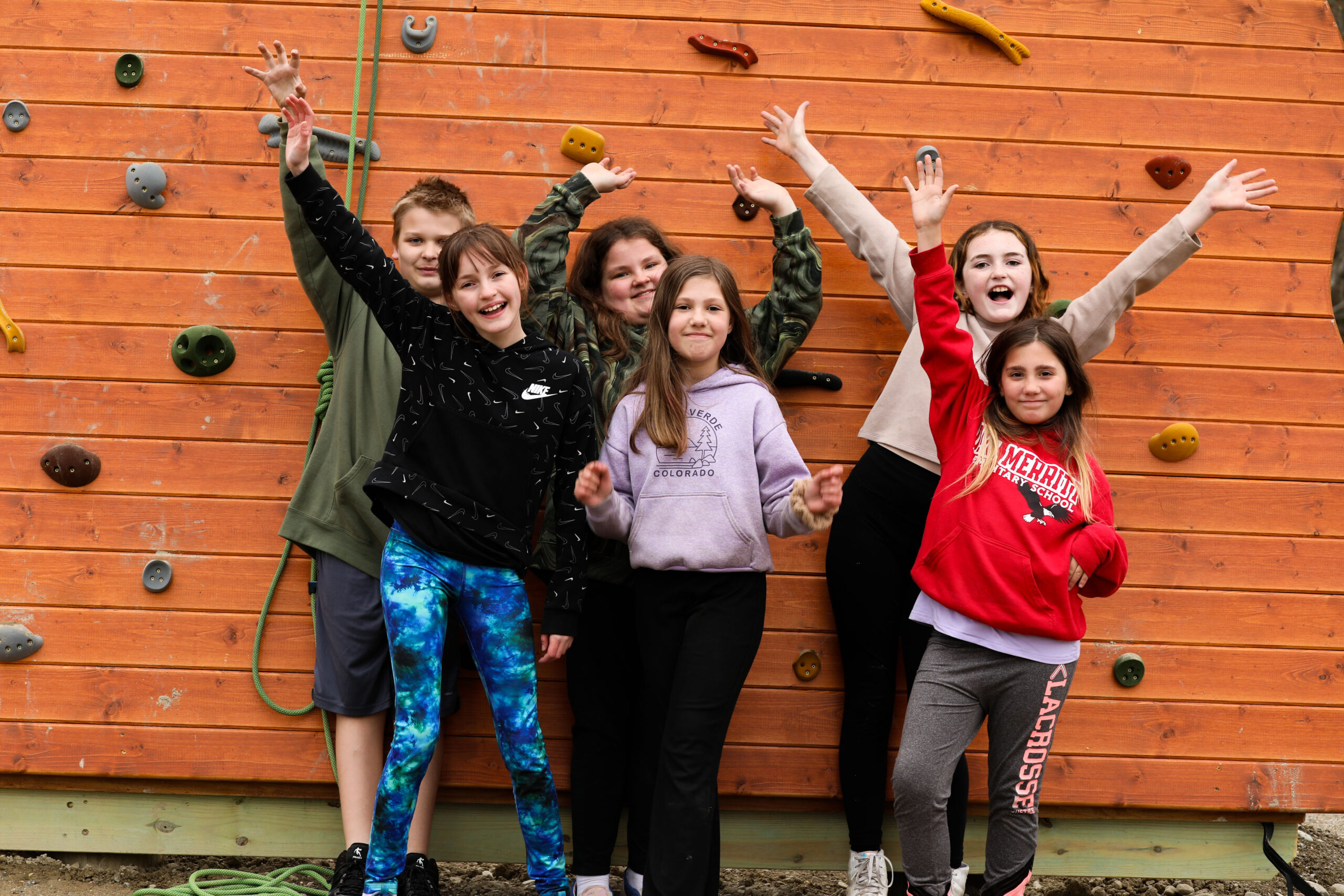 A photo of six students, five girls and one boy, raising their hands and cheering in front of a climbing wall