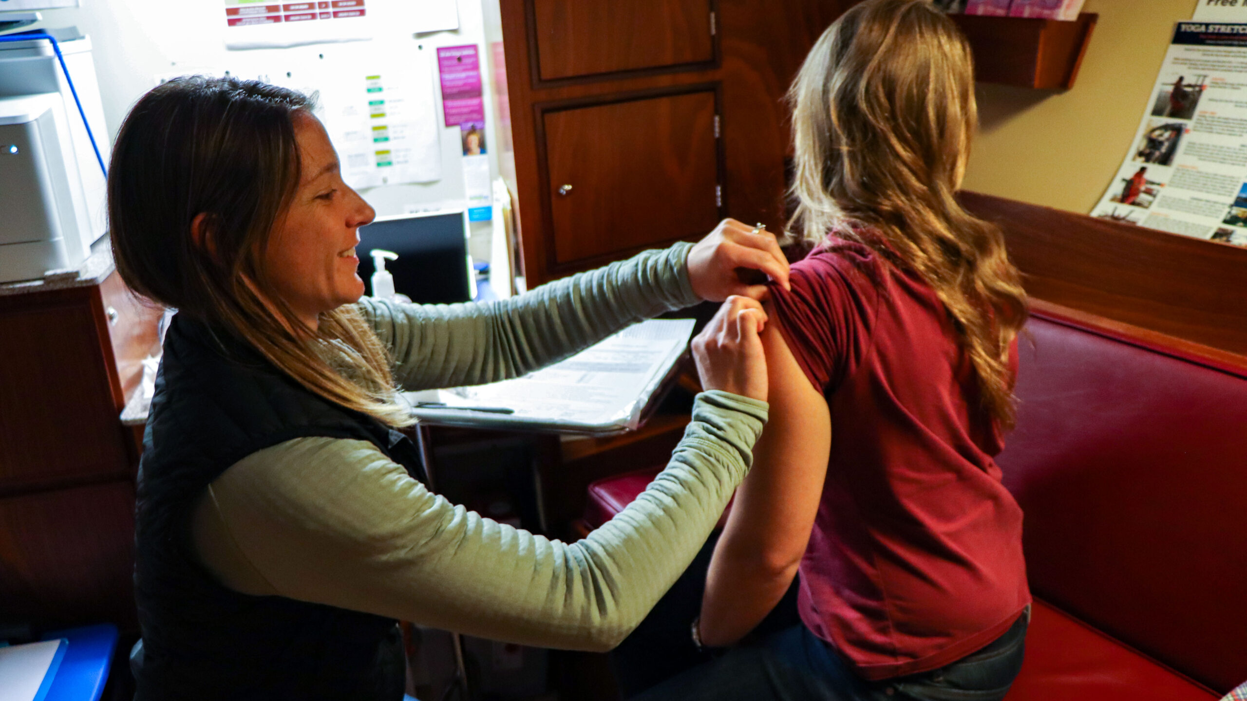 A photo of two young women. One is giving a shot to the other one.